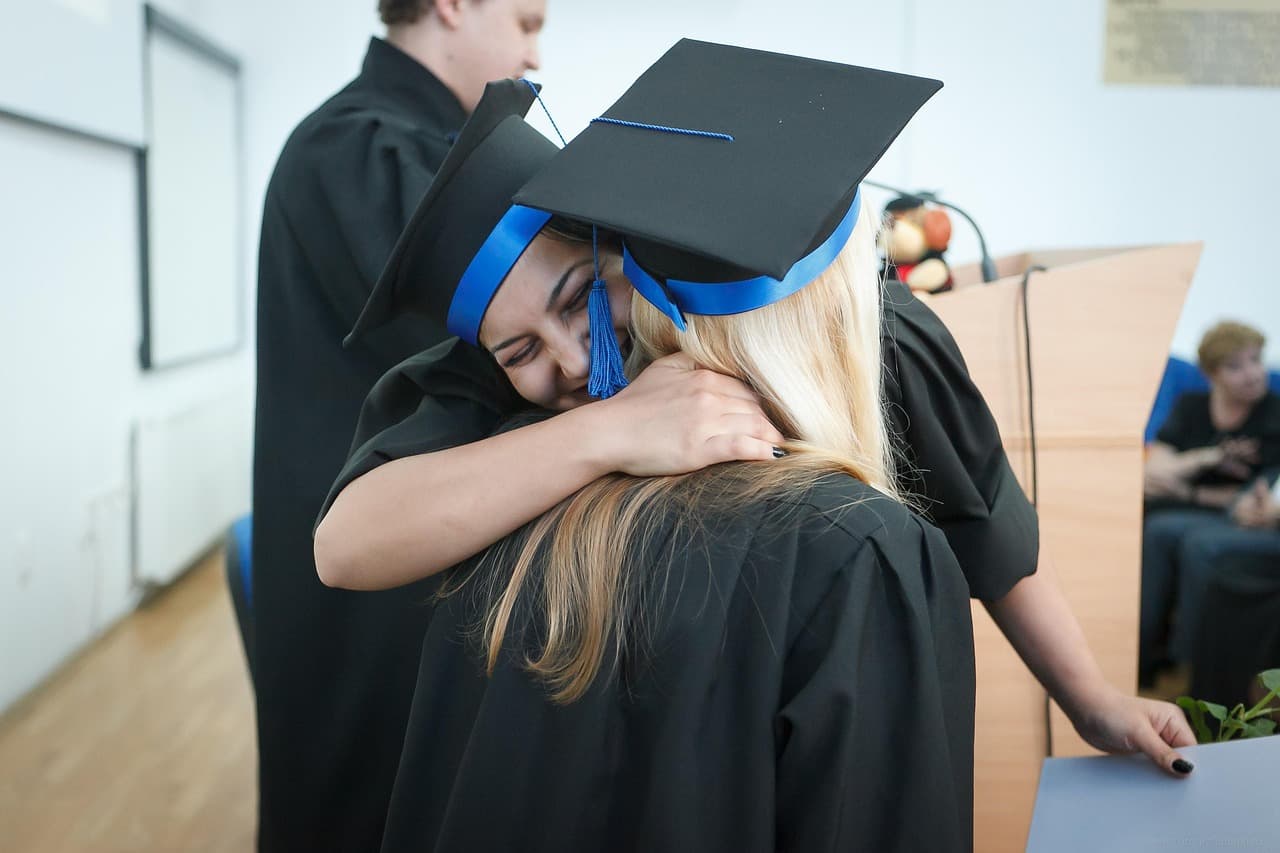 Two graduates in black gowns and caps with blue trim hugging at a graduation ceremony