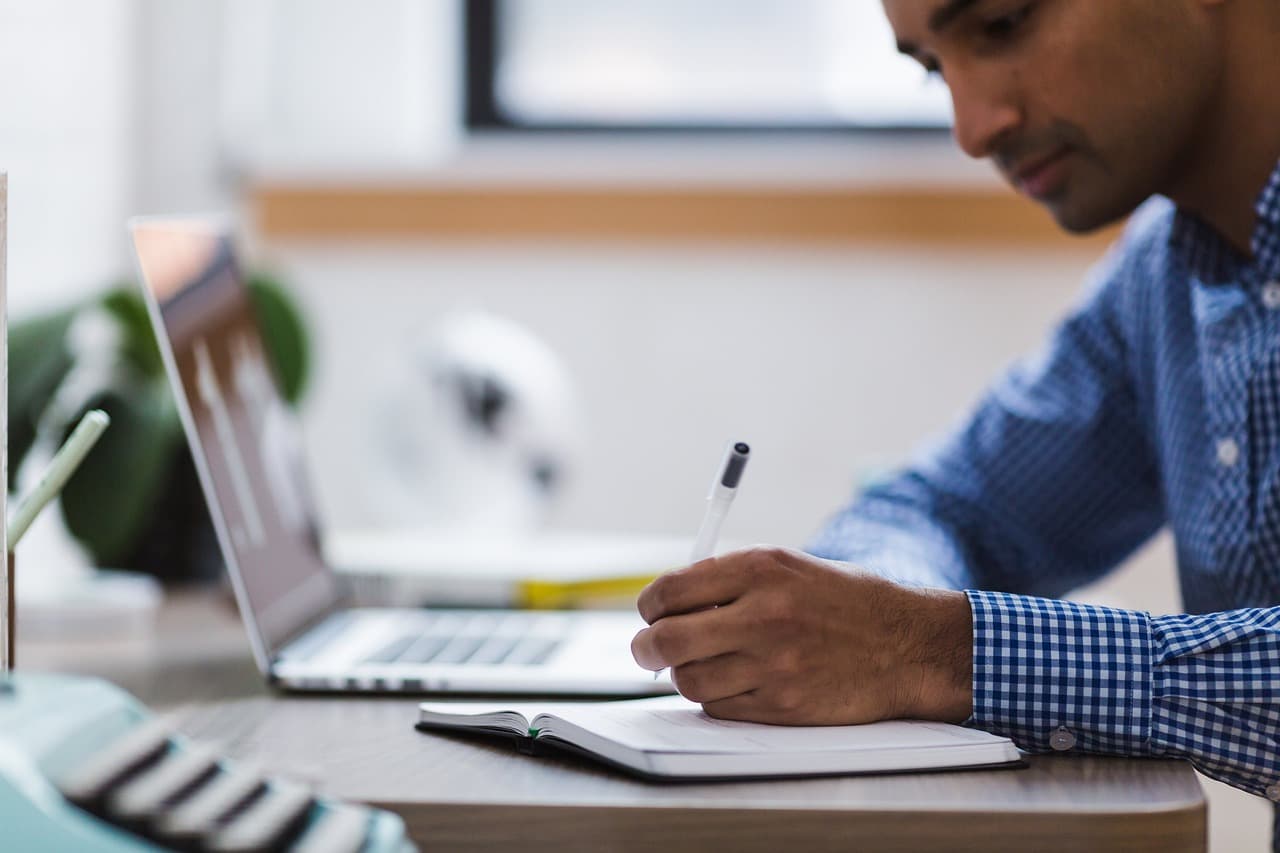 Student writing in a notebook next to a laptop while doing research